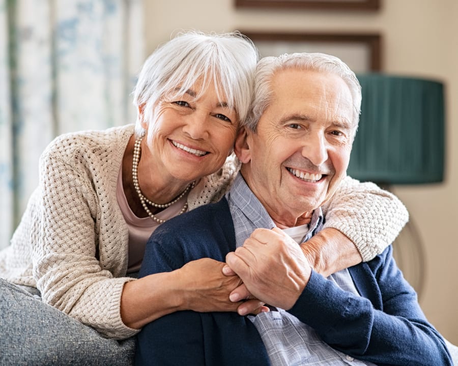 An older man is sitting on a couch while an older woman hugs him from behind.