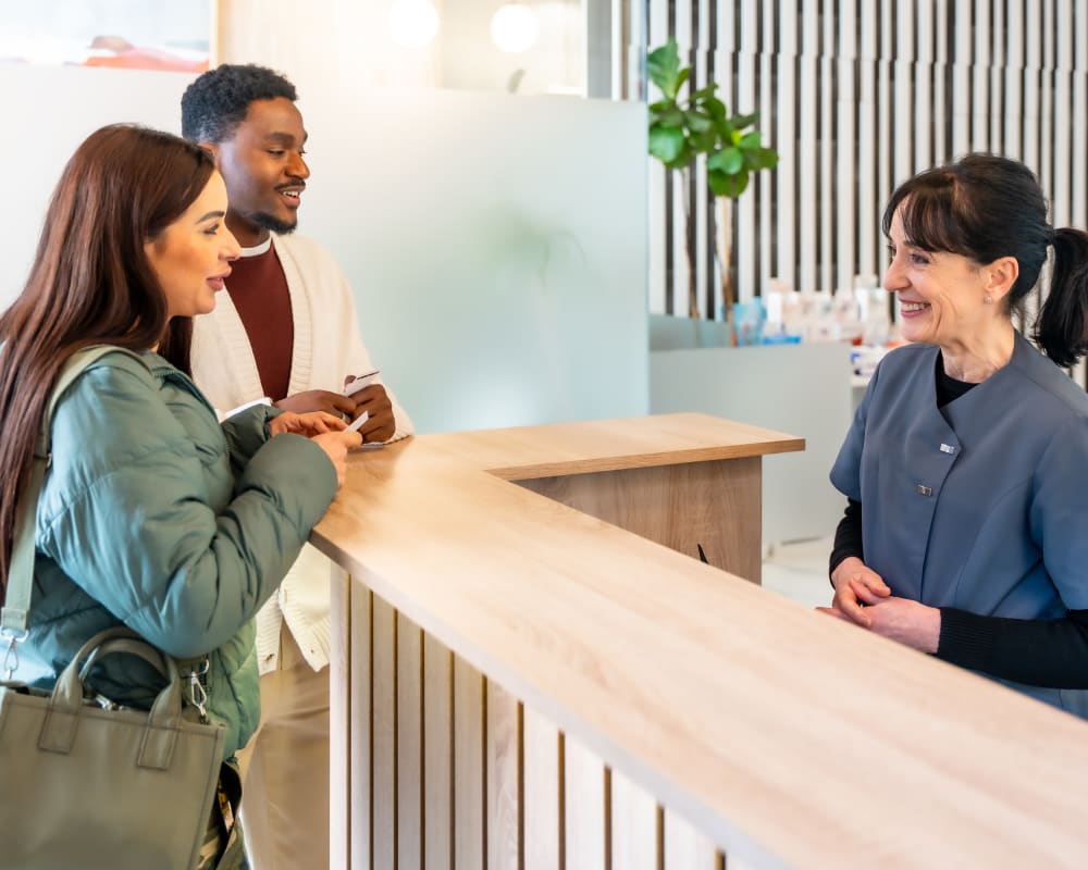 A dental professional is talking to two patients at the office's reception desk. They are leaning on the desk and holding pieces of paper.