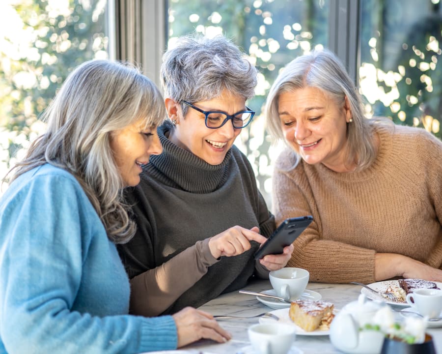 A group of three older women smile as they all look at the woman in the middle's phone. They are sitting together having tea and pastries.