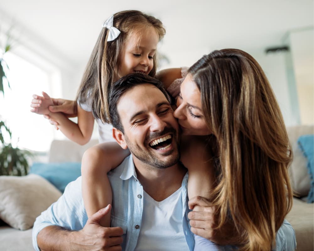 A young girl is sitting on her father's shoulder, and the mom is standing behind them, kissing the dad on the cheek while he smiles.